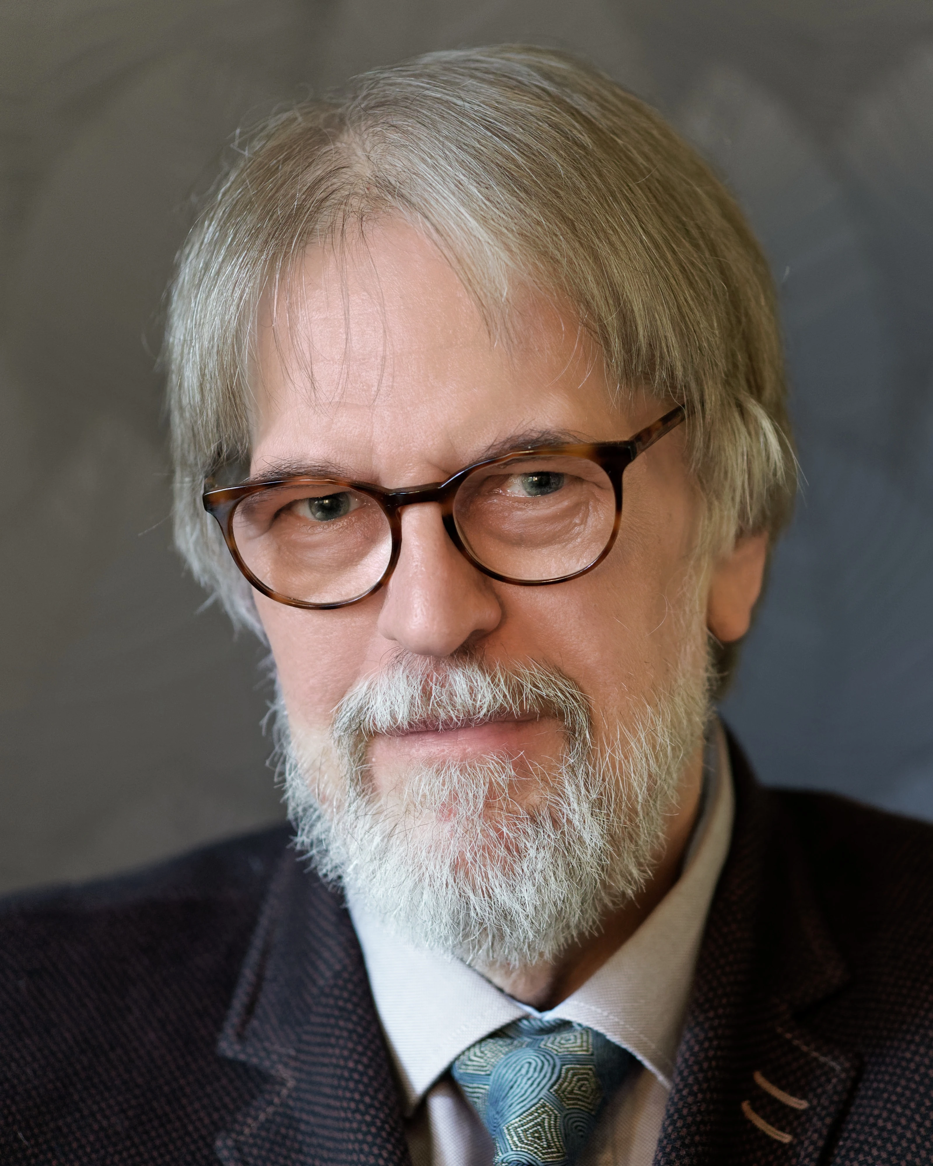 A man in his sixties with glasses and a beard wearing a suit and tie